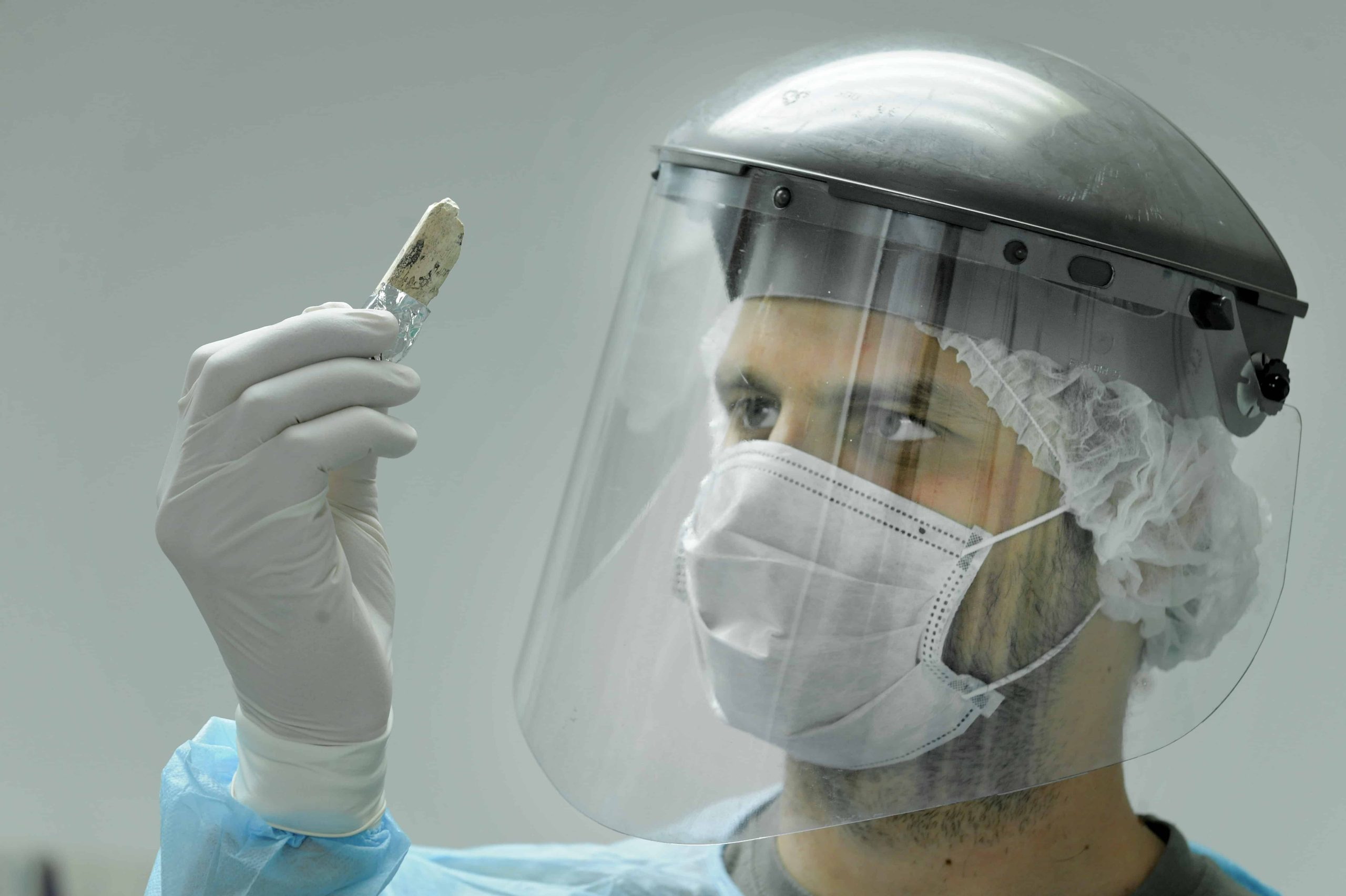 A researcher wearing a visor and face mask examines a bone he is holding in his hand.